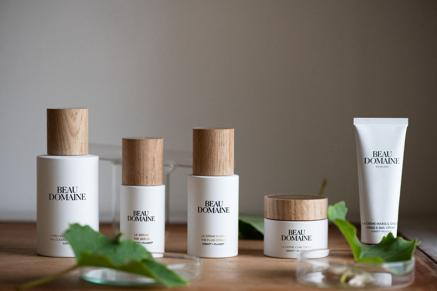 Cinq produits Beau Domaine (L’Émulsion Nettoyante, Le Sérum, La Crème Fluide, La Crème et La Crème Mains & Ongles) disposés en ligne sur une table en bois, avec des feuilles de vigne floues au premier plan et une lumière douce en arrière-plan
