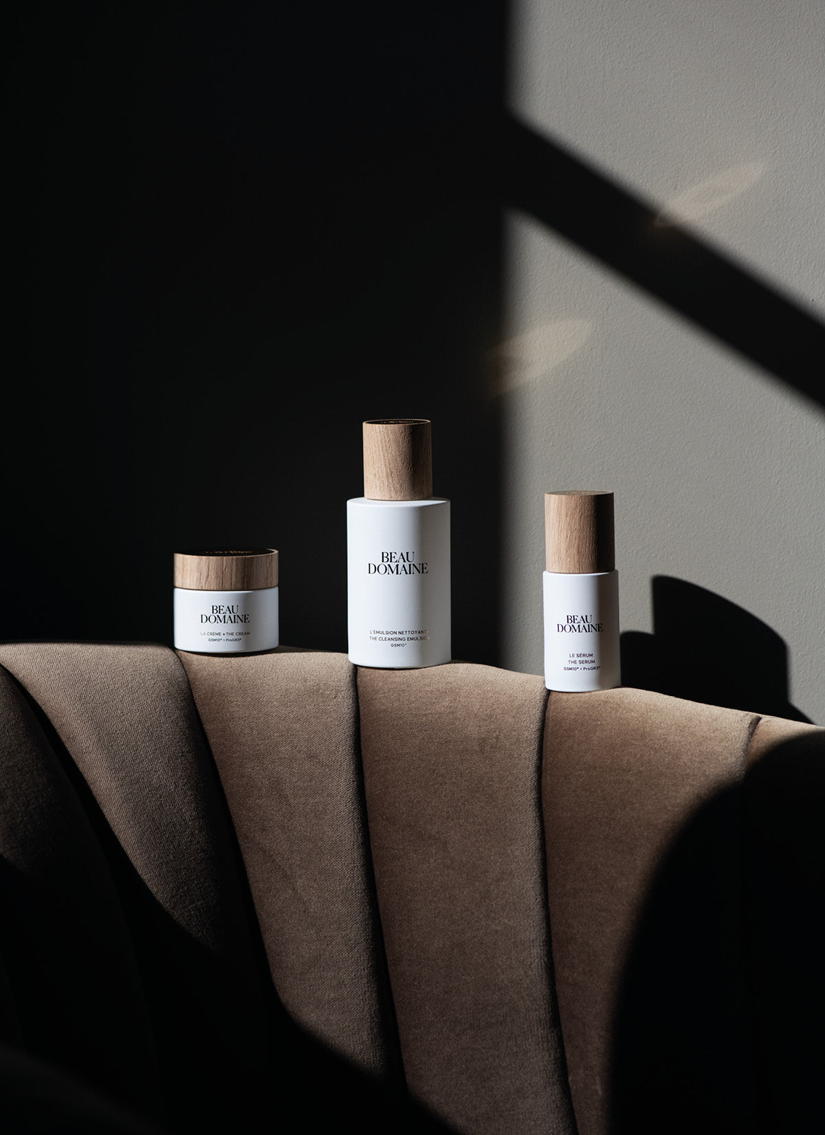 Trois soins Beau Domaine – La Crème, Le Sérum et La Crème Riche – disposés sur un fauteuil en velours, dans une lumière douce avec ombres graphiques sur le mur.